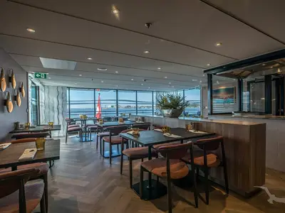 Ein modernes Restaurant an Bord der MS Annabelle mit großen Panoramafenstern zum Wasser. Es gibt dunkle Tische, orangefarbene Stühle und eine offene Küche. Die Tische sind gedeckt.