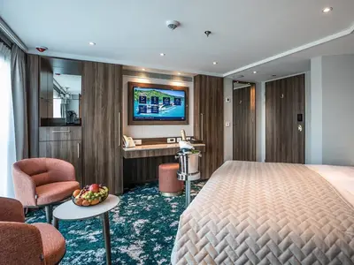 Geräumige Suite an Bord der MS Annabelle mit Doppelbett, Sitzecke, Obstkorb, großem TV und Minibar. Modernes Interieur mit warmen Holztönen und Teppichboden.
