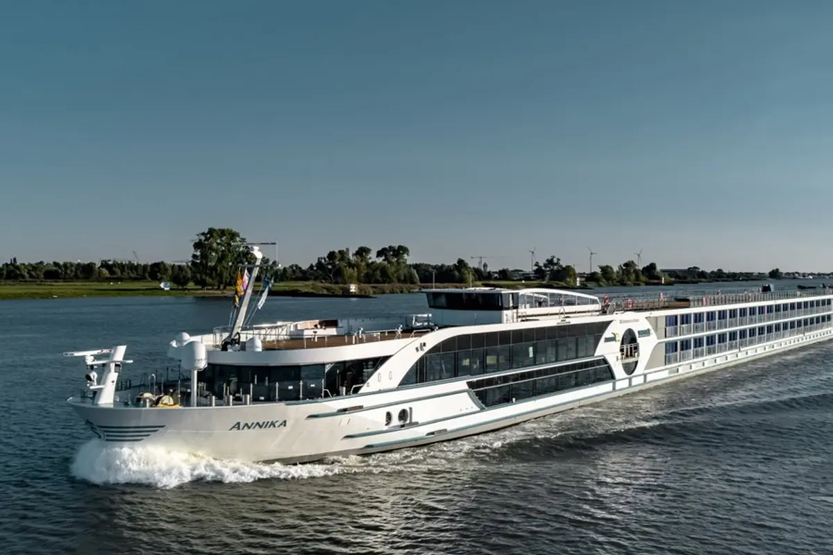 Das moderne Flusskreuzfahrtschiff MS Annika fährt auf einem Fluss. Das weiße Schiff erzeugt eine Bugwelle. Grüne Ufer und Windräder sind unter blauem Himmel sichtbar.