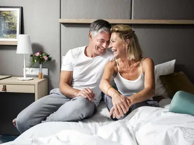 Ein glückliches Paar mittleren Alters lacht und schaut sich liebevoll auf dem Bett einer modern eingerichteten Kabine an Bord der MS Annika an. Sie tragen legere Kleidung.