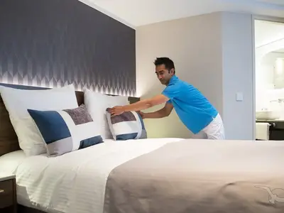 Ein lächelndes Crewmitglied der MS Annika in blauem Shirt macht ein Bett in einer modernen Kabine. Weiße Bettwäsche, blaue Zierkissen und ein Badezimmer sind sichtbar.