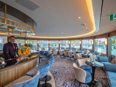 Ein Barkeeper lächelt hinter der Bar der modernen Barlounge an Bord der MS Annika. Große Fenster bieten einen weiten Blick auf den Fluss.
