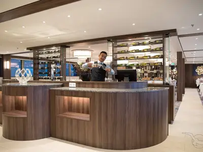 Ein lächelnder Barkeeper in Weste schenkt an einer modernen Bar der MS Annika ein Getränk ein. Im Hintergrund eine beleuchtete Weinwand und gedeckte Tische sind sichtbar.
