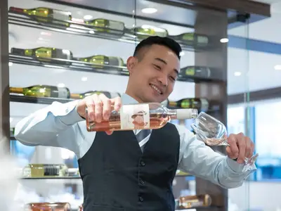 Ein lächelndes Crewmitglied der MS Annika in Weste und Hemd gießt Roséwein aus einer Flasche in ein Glas. Im Hintergrund sind Weinflaschen in einem Regal zu sehen.