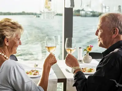 Ein lächelndes älteres Paar toastet mit Weißwein im eleganten Restaurant der MS Annika. Auf dem festlich gedeckten Tisch stehen Speisen, im Hintergrund ist Wasser durch große Fenster sichtbar.