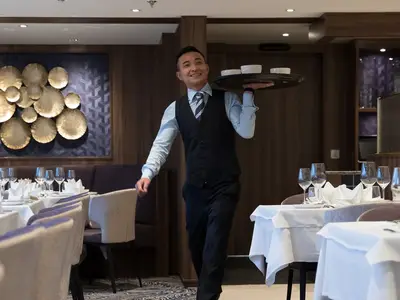 Ein lächelnder Kellner in Uniform trägt eine Servierplatte mit Schalen durch das elegant gedeckte Restaurant der MS Annika. Tische mit weißen Tischdecken und Gläsern sind zu sehen.