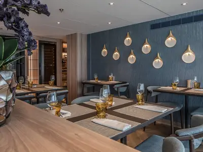 Das elegante Restaurant der MS Annika. Gedeckte Tische mit Gläsern und goldfarbenen Bechern. Blaue Wand mit stilvollen Leuchten. Ein Weinregal im Hintergrund.
