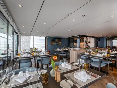 Elegantes Restaurant der MS Annika mit gedeckten Tischen, blauen Stühlen und Holz. Große Fensterfronten bieten Panoramablick auf das Wasser. Ohne Personen.