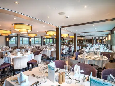 Elegantes Restaurant der MS Annika mit vielen weiß gedeckten Tischen und modernen Stühlen. Warme Deckenleuchten und große Fensterfronten sorgen für eine helle, einladende Atmosphäre.