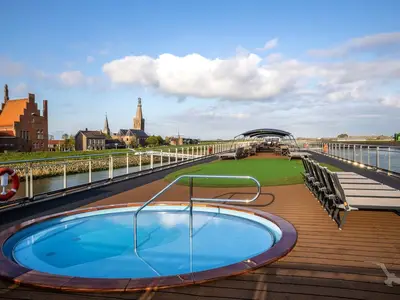 Sonnendeck der MS Annika mit Whirlpool und Liegestühlen. Im Hintergrund Fluss, grüne Ufer und eine europäische Stadt mit Kirchtürmen.