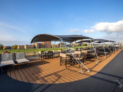 Das Sonnendeck der MS Annika bietet Sitzgelegenheiten unter Sonnensegeln. Mehrere Tische und Stühle laden zum Verweilen ein. Im Hintergrund ist ein Dorf am Flussufer zu sehen.
