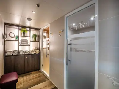 Heller Spa- und Wellnessbereich an Bord der MS Annika. Links ein Regal mit frischen Handtüchern und Dekoration. Rechts sind zwei Glastüren sichtbar, eine davon führt zu einem Dampfbad.