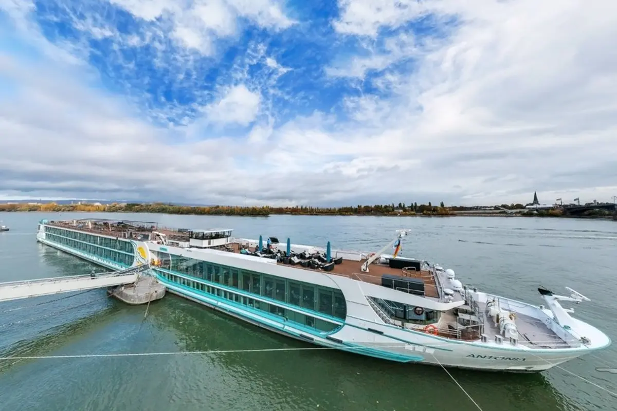 MS Antonia, Flusskreuzfahrtschiff, liegt am Kai. Gangway zum Land. Sonnendeck mit Sitzgruppen, blauen Sonnenschirmen. Fluss, Ufer und bewölkter Himmel.