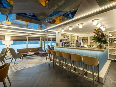 Ein Barmann steht lächelnd hinter der modernen Bar-Theke der MS Antonia. Die Bar-Lounge hat große Fenster mit Blick auf das Wasser und eine helle, elegante Einrichtung.