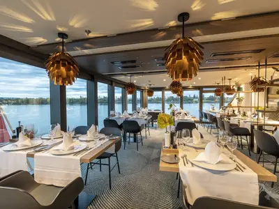 Das elegante Restaurant der MS Antonia zeigt gedeckte Tische, moderne Leuchten und große Fenster mit Blick auf einen ruhigen Fluss mit grünen Ufern.