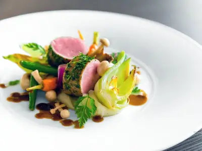 Nahaufnahme eines Gourmet-Gerichts auf einem weißen Teller. Zu sehen ist zartrosa Fleisch mit Kräuterkruste, dazu farbenfrohes Gemüse wie Karotten und grüne Bohnen, Pilze und eine dunkle Soße. Ein elegantes Mahl an Bord der MS Antonia.