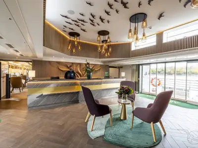 Moderne Rezeption an Bord der MS Antonia. Ein Crew-Mitglied steht hinter dem Schalter aus Marmor und Gold. Im Vordergrund sind Lounge-Sessel und ein runder Teppich zu sehen. Große Fenster bieten Flussblick. Decke mit Vogel-Dekor und Hängelampen.