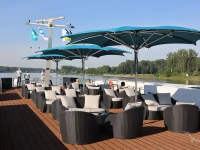 Das Sonnendeck der MS Antonia bietet bequeme Lounge-Sessel unter blauen Sonnenschirmen. Mehrere Passagiere entspannen an Bord der MS Antonia und genießen den Blick auf das bewaldete Flussufer am Tag.
