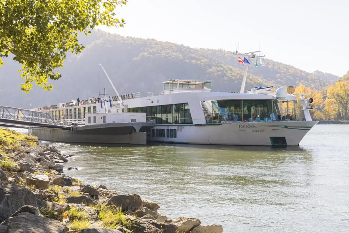 Die weiße MS Ariana liegt an einem felsigen Flussufer. Im Hintergrund erheben sich bewaldete Berge mit Herbstlaub unter hellem Himmel.