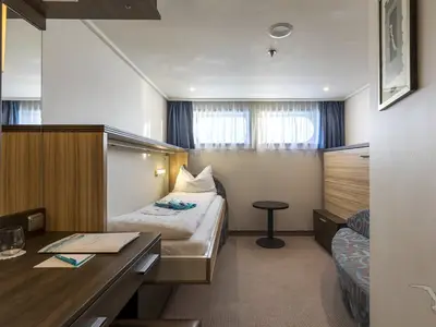 Eine helle Außenkabine an Bord der MS Ariana. Sie zeigt ein Einzelbett mit weißer Bettwäsche, kleine runde Fenster mit Vorhängen, einen dunklen Schreibtisch, einen kleinen Tisch und eine Sitzbank mit Muster. Die Kabine ist freundlich und funktional eingerichtet mit Holzmöbeln.
