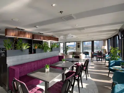 Moderne Lido Bar an Bord der MS Ariana mit lila Bankett, teal Sesseln und Holztischen. Große Fenster bieten Flussblick und Stadtpanorama. Grüne Pflanzen dekorieren den Raum, der zum Entspannen einlädt.