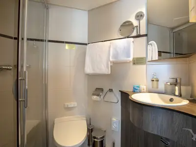 Helles Badezimmer an Bord der MS Ariana mit weißen Fliesen, verglaster Duschkabine, WC, einem runden Waschbecken und einem Spiegel. Frische Handtücher hängen bereit.