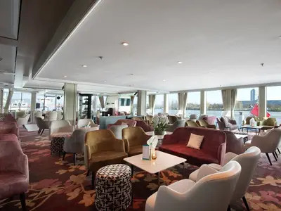 Eine geräumige, elegant eingerichtete Lounge an Bord der MS Ariana mit großen Panoramafenstern. Verschiedene Sitzgruppen in warmen Farbtönen und ein Blick auf das Flussufer sind zu sehen.