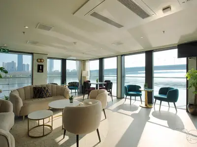 Eine helle, moderne Lounge an Bord der MS Ariana mit großen Panoramafenstern, die einen weiten Blick auf einen Fluss und eine städtische Skyline bieten. Verschiedene Sitzgruppen laden zum Verweilen ein.