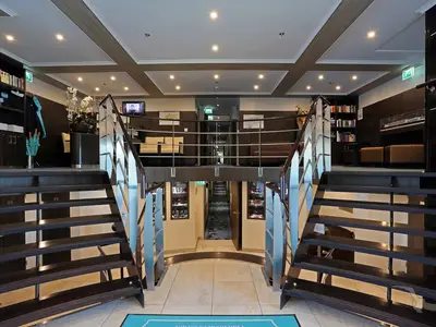 Blick in das elegant gestaltete Foyer der MS Ariana mit dunklen Holztreppen, einem Barbereich, Sitzgelegenheiten und einer kleinen Bibliothek.