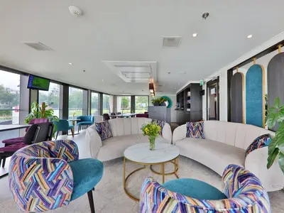 Moderne Lounge an Bord der MS Ariana mit hellen Sofas, farbigen Sesseln und großem Couchtisch. Fenster bieten Flussblick. Entspanntes Ambiente mit Pflanzen.