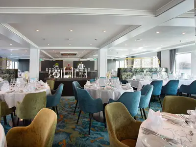 Das elegante Restaurant der MS Ariana ist mit runden Tischen, teal- und olivfarbenen Stühlen und einem Buffetbereich ausgestattet. Große Fenster bieten einen Blick auf die Flusslandschaft.