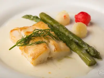 Eine gebratene Alaska Heilbuttschnitte in cremiger Trüffel Beurre Blanc, garniert mit frischem Dill. Daneben grüne Spargelstangen und Kartoffelperlen, ein elegantes Gericht an Bord der MS Ariana.