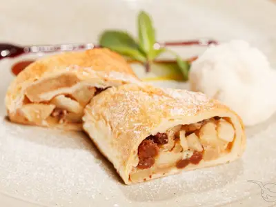 Nahaufnahme eines frisch gebackenen, leicht gezuckerten Apfelstrudels, mit Rosinen und Apfelstücken gefüllt, serviert mit einer Kugel Eiscreme auf einem Teller an Bord der MS Ariana.