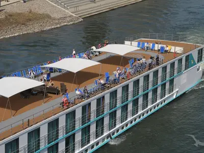 Luftaufnahme der MS Ariana, die auf einem Fluss fährt. Gäste entspannen auf blau-weißen Liegestühlen unter weißen Sonnensegeln auf dem oberen Deck. Der Fluss und felsiges Ufer sind sichtbar.