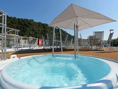 Ein Jacuzzi mit blauem Wasser auf dem Sonnendeck der MS Ariana unter einem Sonnenschirm. Im Hintergrund bequeme Stühle und grüne bewaldete Berge.