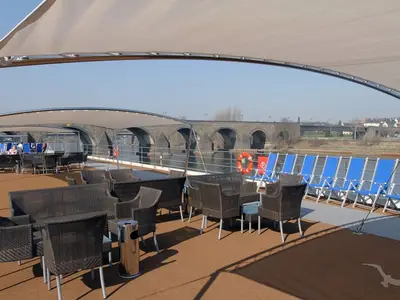 Einladendes Sonnendeck der MS Ariana mit Rattanmöbeln, Liegestühlen und Sonnenschutz, dahinter eine historische Steinbrücke über einen Fluss.
