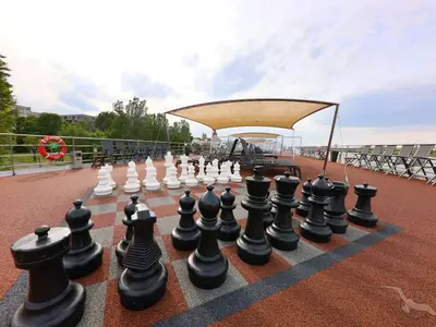Ein riesiges Schachspiel mit schwarzen und weißen Figuren auf dem Sonnendeck der MS Ariana. Im Hintergrund sind Liegestühle, Sonnensegel und eine Uferpromenade zu sehen. Der Himmel ist bewölkt.