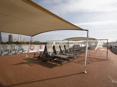 Das Sonnendeck der MS Ariana mit beigen Sonnensegeln und zahlreichen grauen Liegestühlen. Im Hintergrund ist eine Stadtkulisse an einem Fluss zu sehen. Ein Rettungsring ist an der Reling befestigt.