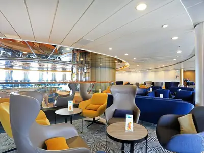 Blick in Harry's Bar an Bord der MS Artania. Die Lounge ist mit verschiedenen blauen Sofas und gelben sowie grauen Sesseln ausgestattet. Auf den Tischen stehen bunte Drinks. Große Fenster bieten Tageslicht. Das Design umfasst spiegelnde Oberflächen. Die MS Artania bietet eine moderne und gemütliche Atmosphäre.