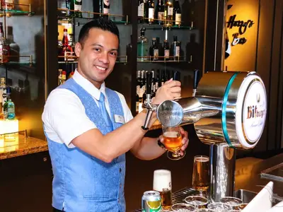 Ein lächelnder Barmann in blauer Weste zapft ein Glas Bitburger Bier an Bord der MS Artania. Die gut ausgestattete Harry's Bar mit Spirituosenregalen ist sichtbar.
