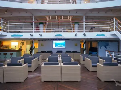 Blick in die helle Phoenix Bar auf der MS Artania mit gemütlichen Korbstühlen und dunkelblauen Kissen. Im Hintergrund eine Bar und ein großer Bildschirm.