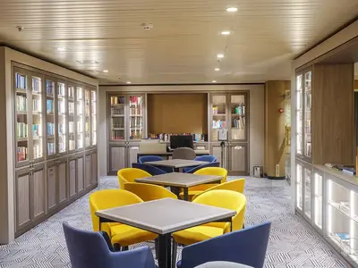 Geräumige Bibliothek an Bord der MS Artania mit Bücherregalen und Lesebereichen. Mehrere Tische mit leuchtend gelben und blauen Sesseln laden zum Verweilen ein.