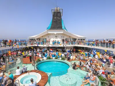Weitwinkelaufnahme vom Sonnendeck der MS Artania mit zahlreichen Gästen, die sich um den Pool, die Kopernikus Bar und Sitzbereiche versammeln. Sonniges Wetter, offenes Meer im Hintergrund.