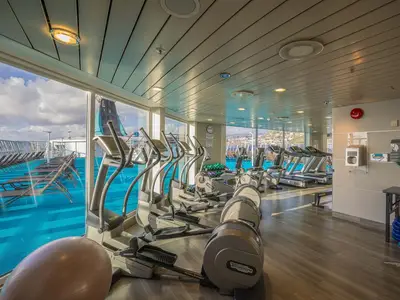 Ein modernes Fitnessstudio an Bord der MS Artania mit einer Reihe von Cardiogeräten und Hanteln, die vor großen Fenstern mit Blick auf das Meer und ein Pooldeck stehen.