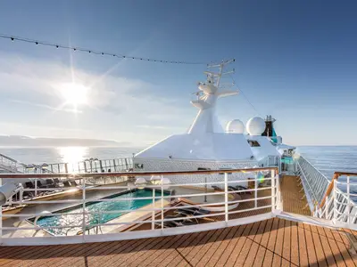 Blick über den Poolbereich auf dem Sonnendeck der MS Artania. Das glitzernde Meer und der Schiffsaufbau unter strahlendem Sonnenschein. Liegestühle um den Pool sind zu sehen.