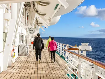 Eine Gruppe von drei Personen spaziert bei blauem Himmel und Sonnenschein auf dem Promenadendeck der MS Artania. Die offene See ist rechts sichtbar.
