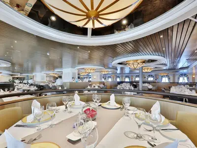 Das elegante Restaurant Vier Jahreszeiten der MS Artania mit festlich gedeckten Tischen, Gläsern und Geschirr. Crewmitglieder im Hintergrund bereiten den Service vor. Der Raum ist stilvoll eingerichtet.