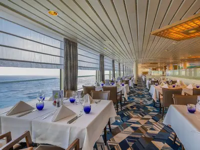 Heller Innenraum des Lido Restaurants an Bord der MS Artania mit stilvoll gedeckten Tischen. Große Fensterfronten bieten einen weiten Blick auf das blaue Meer.