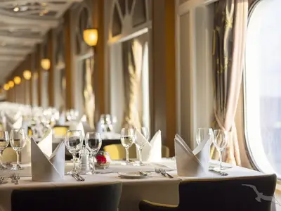 Das helle Restaurant der MS Artania zeigt elegant gedeckte Tische mit weißen Servietten, Gläsern und Besteck. Tageslicht fällt durch die großen Fenster an der Seite ein.