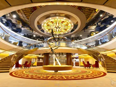 Blick ins elegante Atrium der MS Artania mit geschwungener Treppe, moderner Skulptur, Sitzgelegenheiten und beleuchteter Buntglaskuppel.
